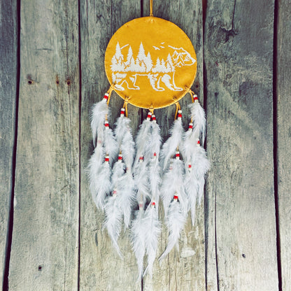 Un capteur de rêves d'inspiration autochtone accroché sur un mur de bois rustique. Le cercle central est en cuir jaune ocre, orné d'une illustration blanche représentant la silhouette d'un ours fusionnant avec une forêt de pins et des montagnes. Plusieurs rangées de plumes blanches duveteuses, agrémentées de petites perles rouges et blanches, pendent élégamment à la base.