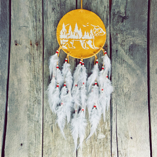 Un capteur de rêves d'inspiration autochtone accroché sur un mur de bois rustique. Le cercle central est en cuir jaune ocre, orné d'une illustration blanche représentant la silhouette d'un ours fusionnant avec une forêt de pins et des montagnes. Plusieurs rangées de plumes blanches duveteuses, agrémentées de petites perles rouges et blanches, pendent élégamment à la base.