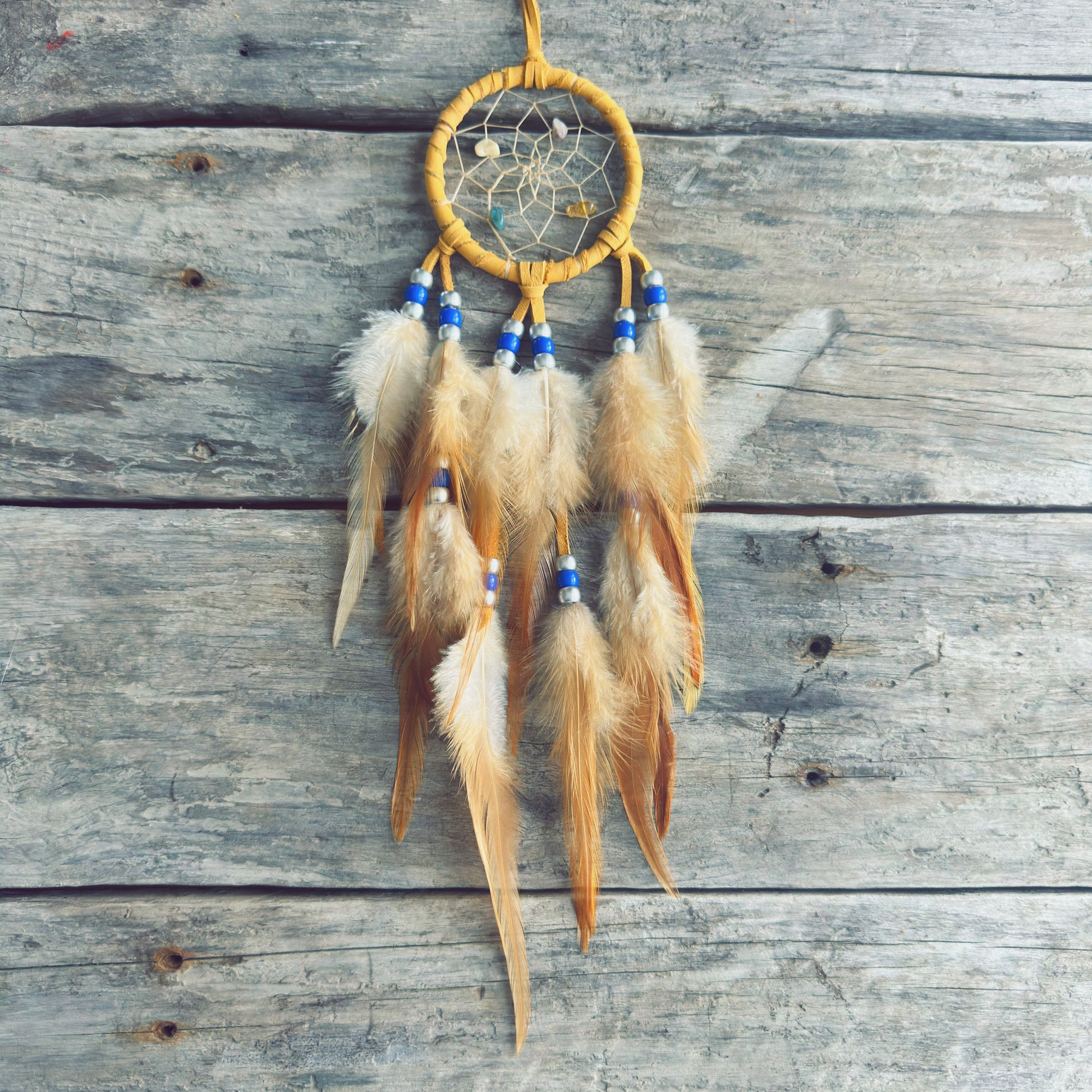 Artisanat autochtone : capteur de rêves de 3 pouces en cuir ocre jaune avec perles bleu royal et blanches, plumes naturelles beige crème sur fond de bois gris