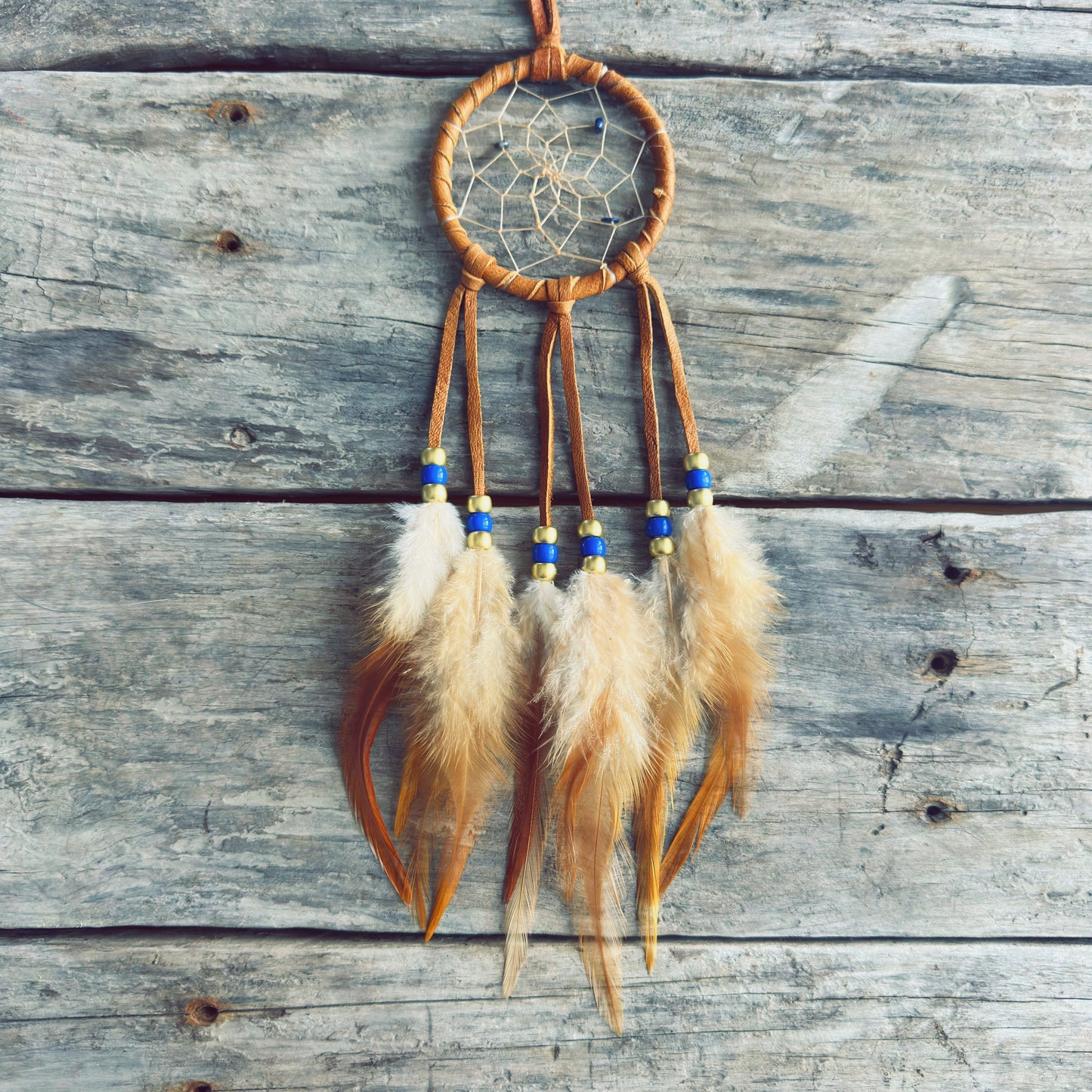 Artisanat autochtone : capteur de rêves de 3 pouces en cuir ocre jaune avec perles bleu royal et dorées, plumes naturelles tachetées sur fond de bois gris