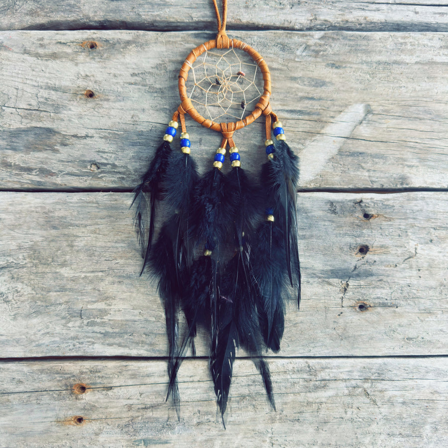 Artisanat autochtone : capteur de rêves de 3 pouces en cuir ocre jaune avec perles bleu royal et dorées, longues plumes noires sur fond de bois de grange