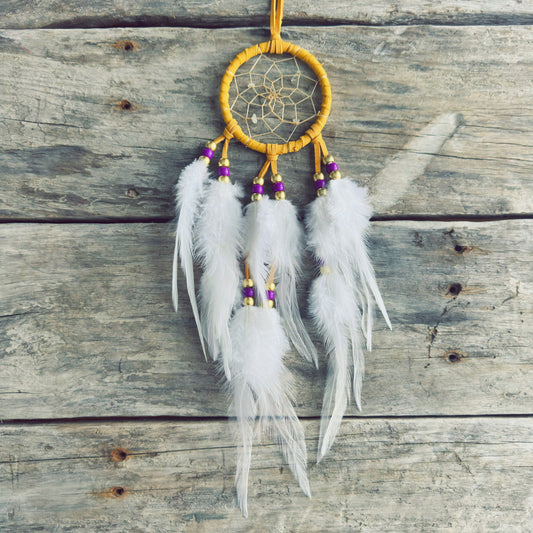 Artisanat autochtone : capteur de rêves de 3 pouces en cuir ocre jaune avec perles mauves et or, plumes blanches vaporeuses sur fond de bois gris.