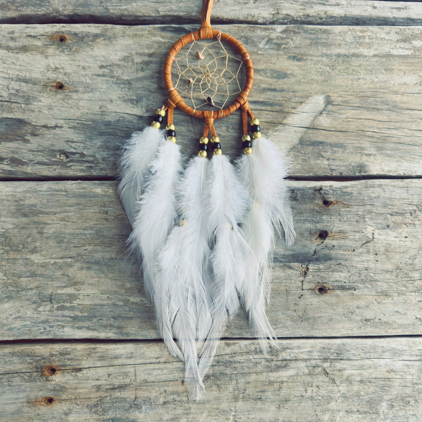Artisanat autochtone : capteur de rêves de 3 pouces en cuir ocre jaune avec perles noires et or, plumes blanches vaporeuses sur fond de bois gris.