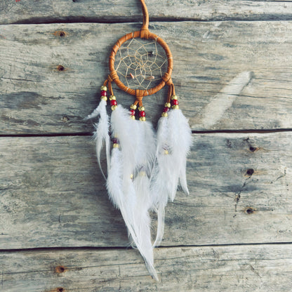 Artisanat autochtone : capteur de rêves de 3 pouces en cuir ocre jaune avec perles rouges et dorées, plumes blanches vaporeuses sur fond de bois gris.