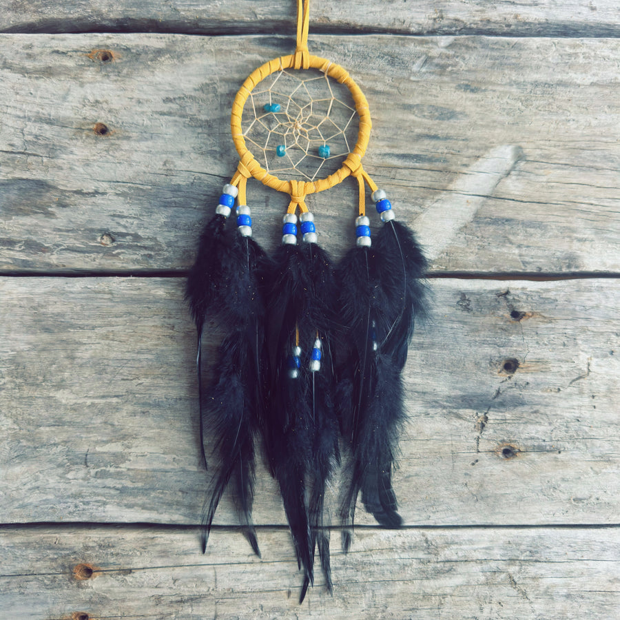 Artisanat autochtone : capteur de rêves de 3 pouces en cuir ocre jaune, perles bleu royal et blanches, avec de longues plumes noires sur fond de bois rustique.