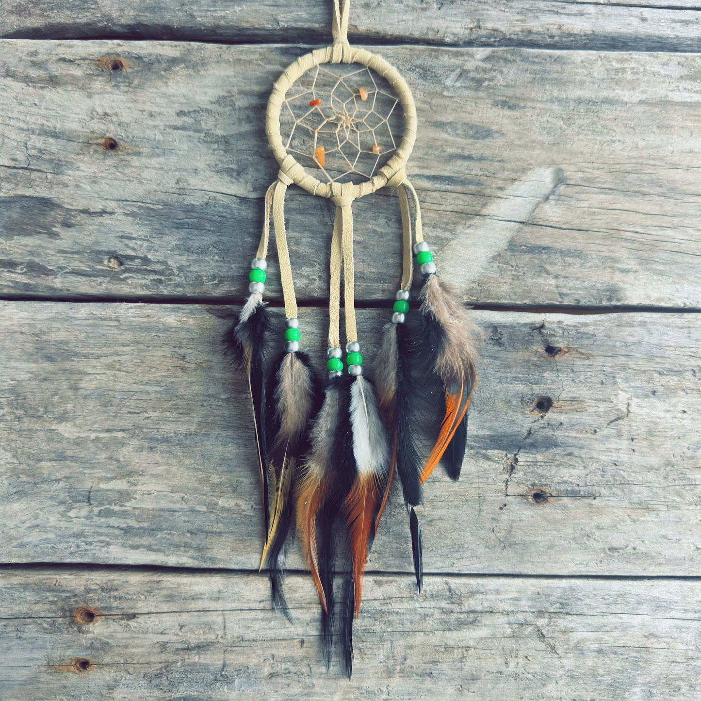 Artisanat autochtone : capteur de rêves de 3 pouces en cuir beige avec perles vert lime et plumes naturelles brunes et orangées sur fond de bois de grange.